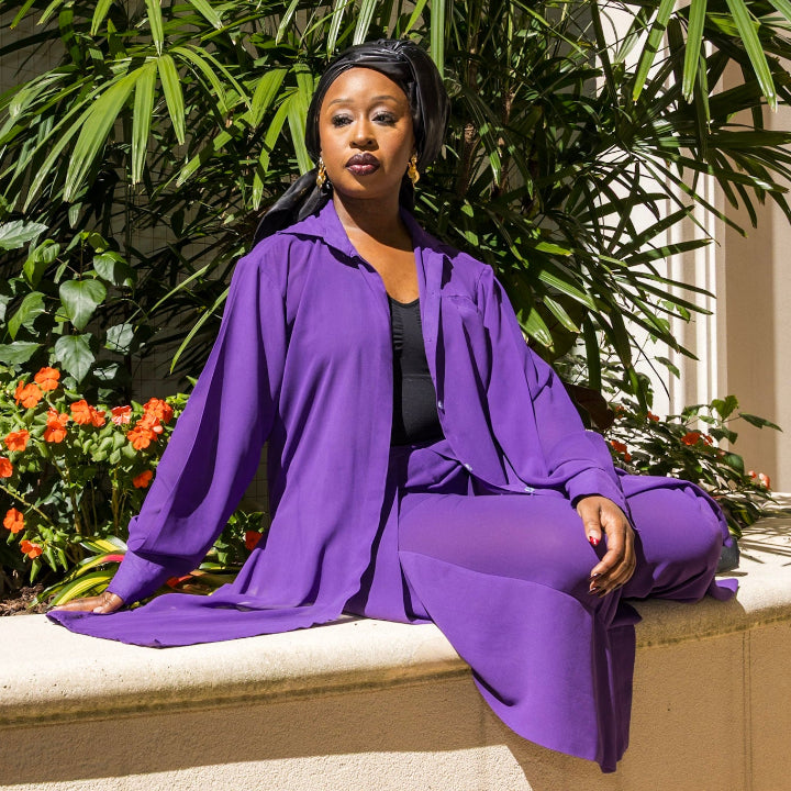 A woman wearing a Matching Set- Kaftan Top, showcasing a vibrant purple matching kaftan set African outfit in a garden setting. This elegant african two piece set features a sheer kaftan shirt and stylish pants, highlighting the beauty of african women attire. The outfit reflects traditional african suit styling and embodies the essence of west african fashion, making it a perfect choice for various occasions. Explore the charm of african kaftan dresses for ladies with this stunning look.